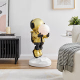 Golden astronaut sculpture holding a lighted moon, placed in a modern living room. The astronaut is standing on a white, faceted base next to a black sideboard and a white armchair.