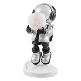 Full view of a silver astronaut sculpture holding a lighted moon. The astronaut is wearing a silver helmet, jacket, and boots, with black gloves and pants, standing on a white, faceted base.