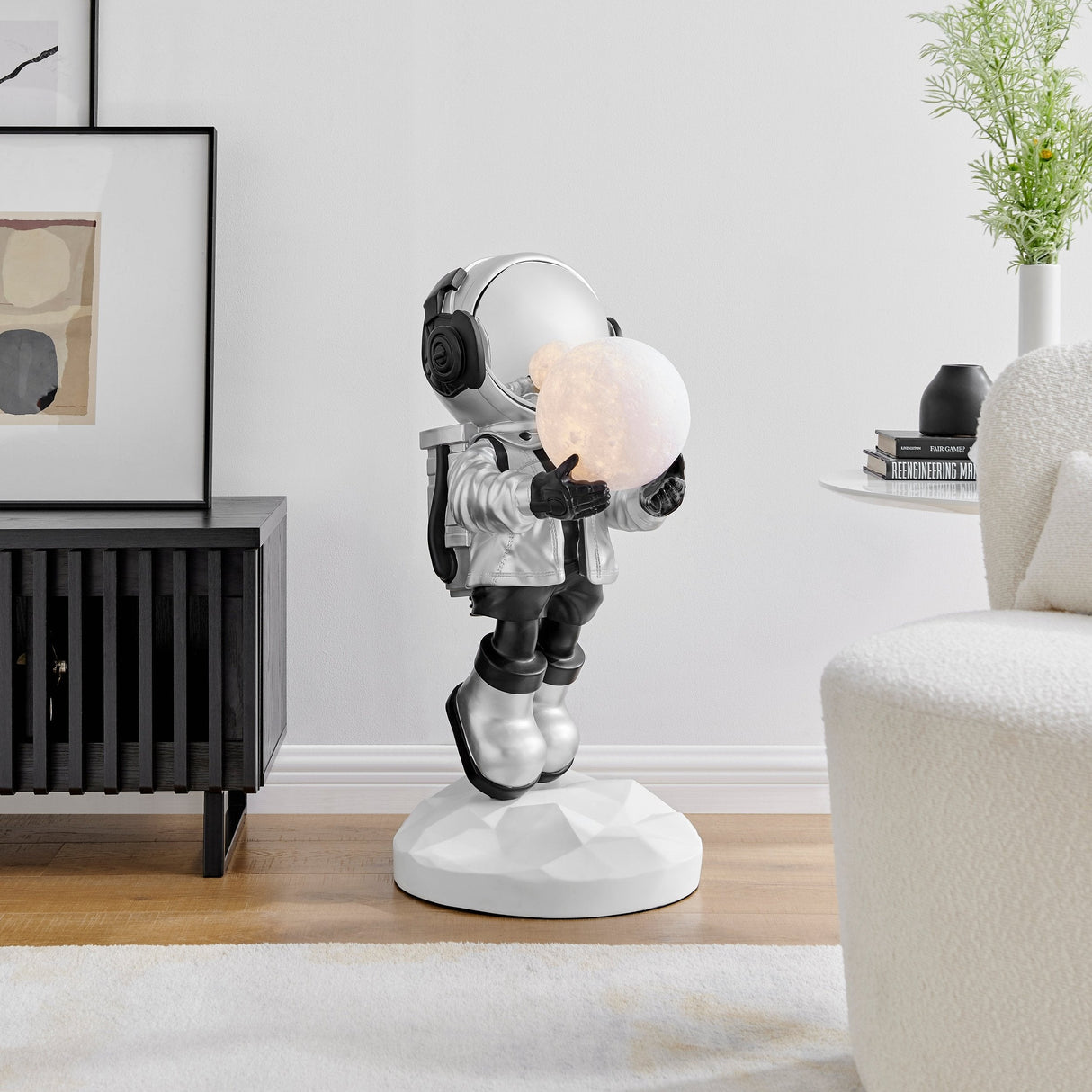  Silver astronaut sculpture holding a lighted moon, placed in a modern living room. The astronaut is standing on a white, faceted base next to a black sideboard and a white armchair.